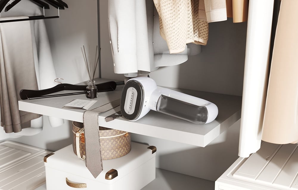 A white steamer stands on a white shelf in a dressing room between hanging clothes and a fragrance diffuser