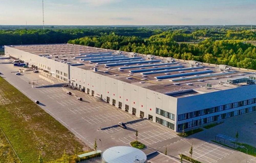A sprawling warehouse building with a flat roof and an adjacent parking lot surrounded by greenery