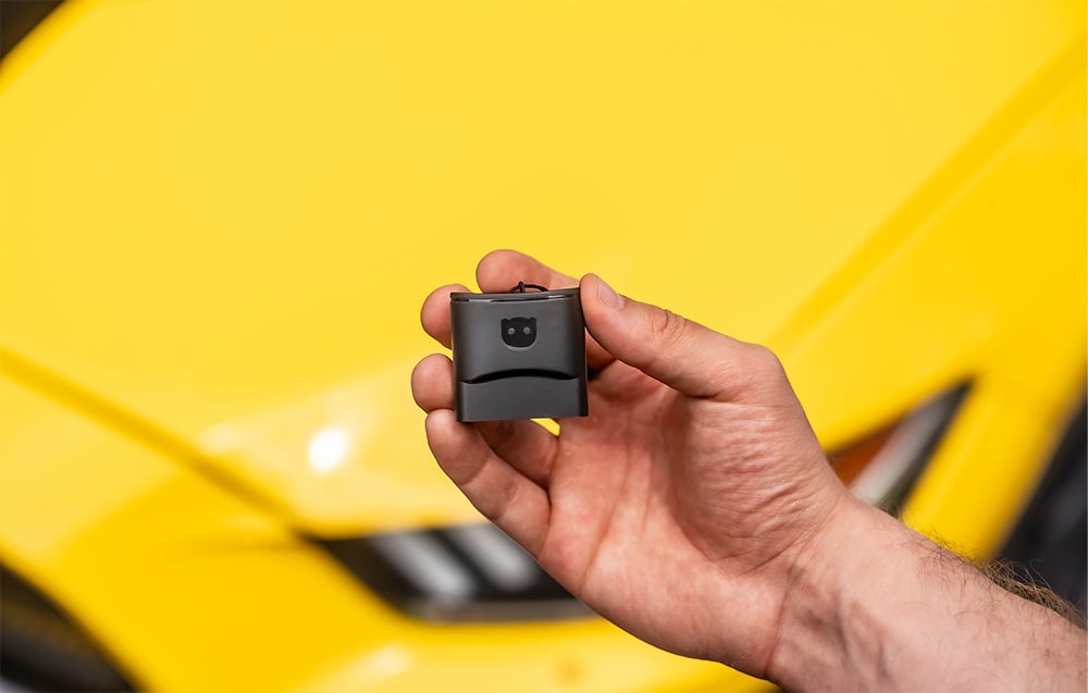 A hand displaying the Carly OBD2 compact adapter against the background of a yellow sports car