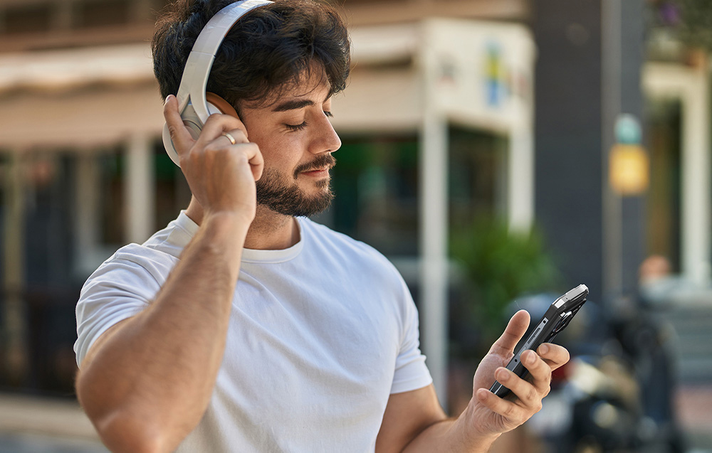 A man wearing white headphones using the HOTWAV smartphone in an urban setting.
