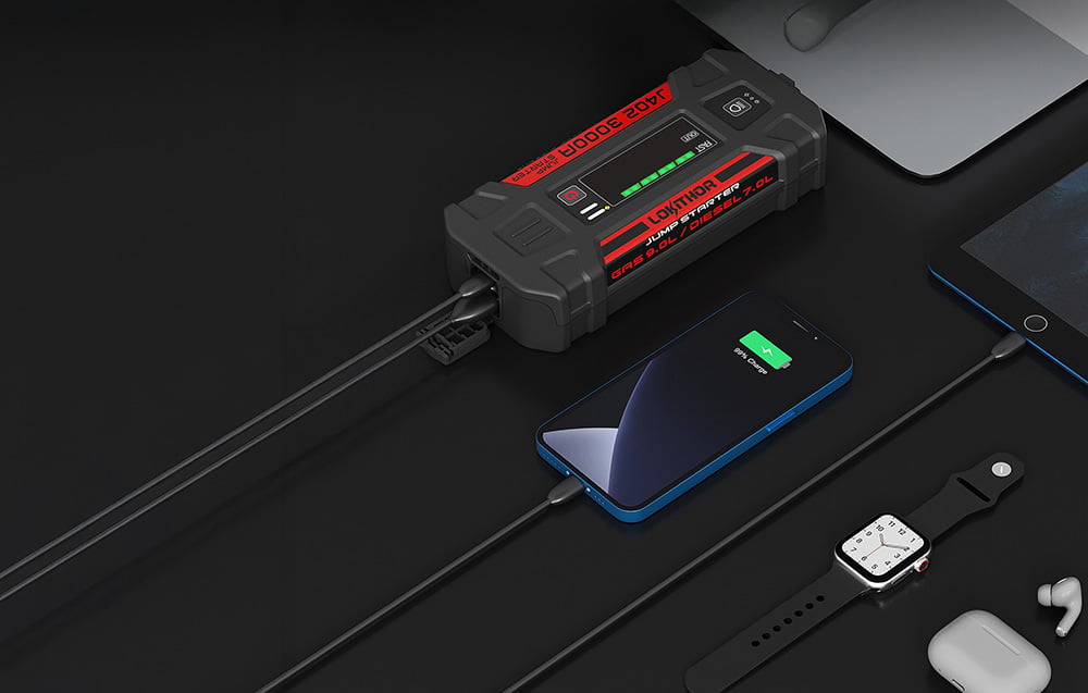 The jump starter charging a phone, watch, and headphones lying on a black tabletop next to a computer.