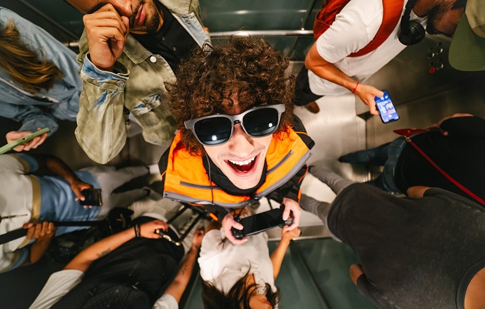 A young man in an orange jacket and sunglasses smiles broadly while standing in a crowded elevator