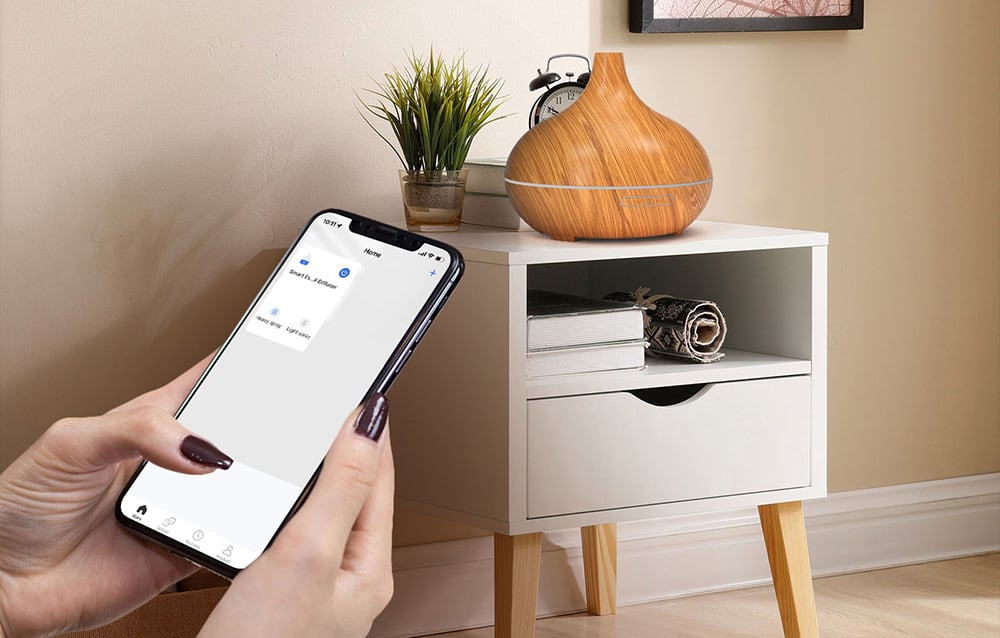 A hand holds a smartphone with an app to control the diffuser, which sits on a white nightstand