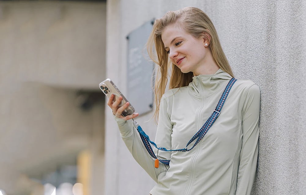 A woman leaning against a wall and using her smartphone, which is mounted to a neck strap