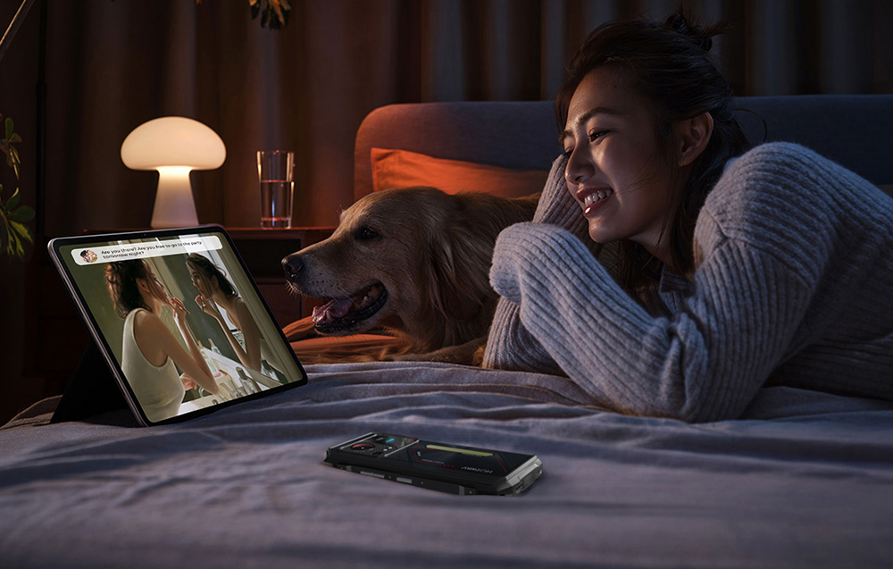 A woman lying on a bed with her dog, watching a video on a tablet; next to her, on the duvet, lies a HOTVWAV smartphone.