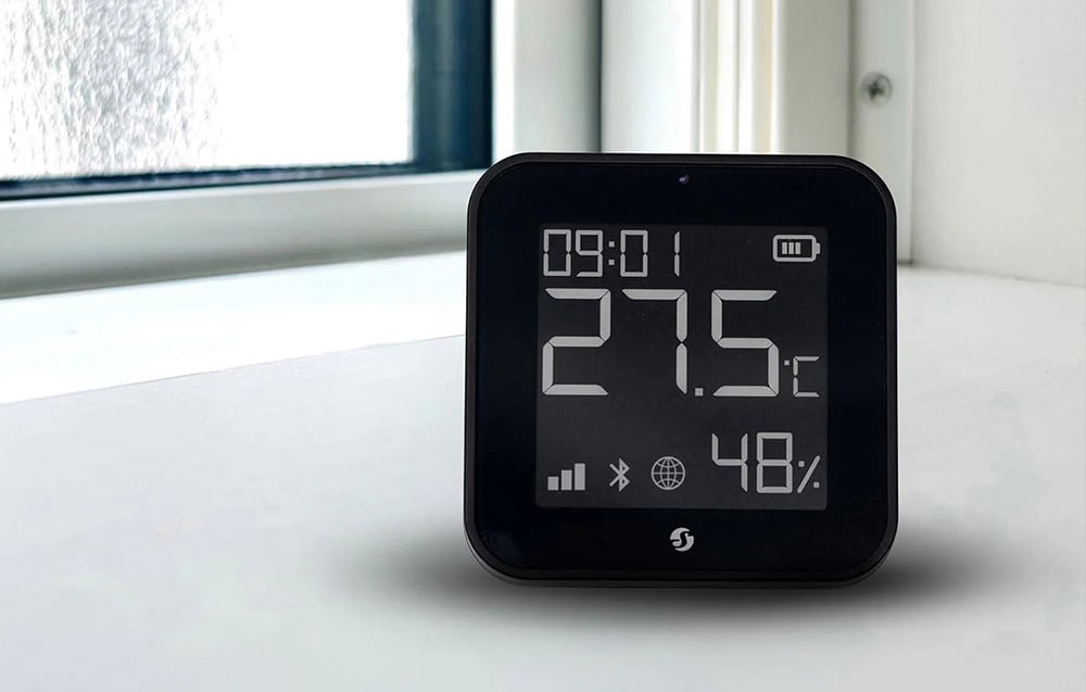 A black room temperature sensor with a display standing on a white windowsill
