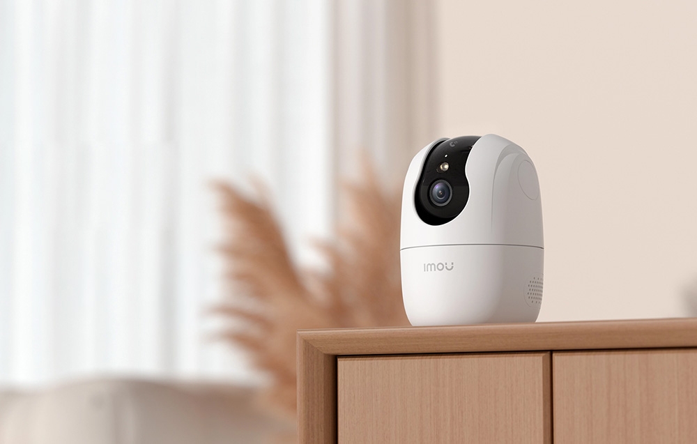 A white Imou camera standing on the edge of a wooden cabinet against a blurred background of the living room