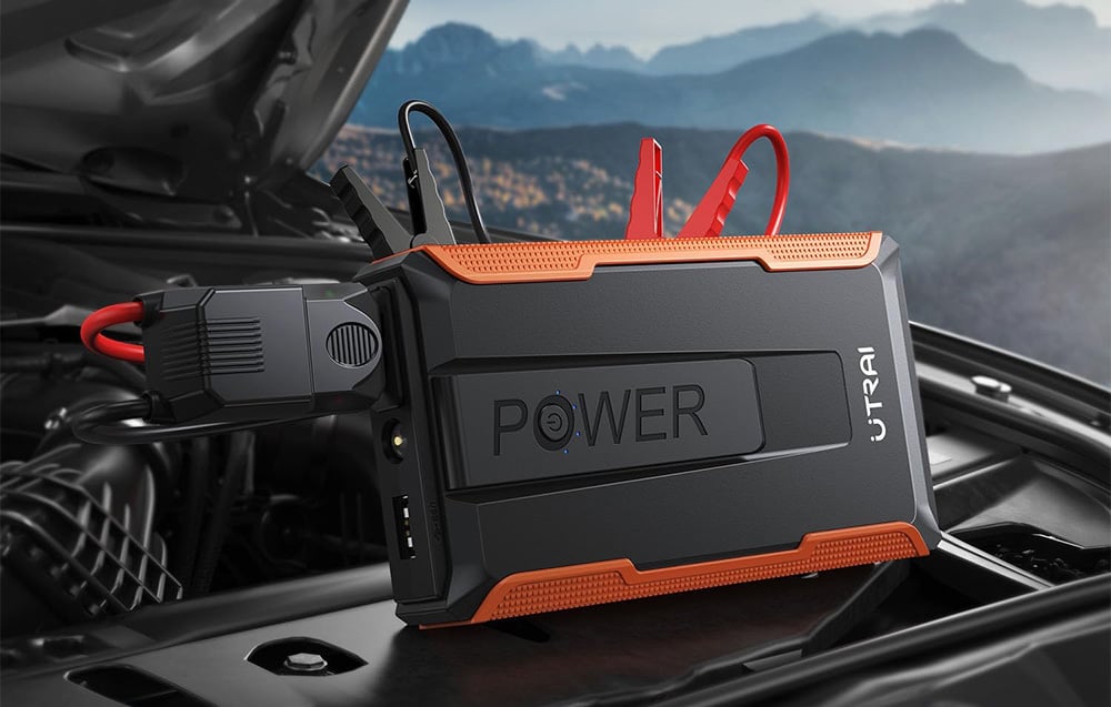 A black car jump starter with orange accents and connected cables rests on the car’s engine.