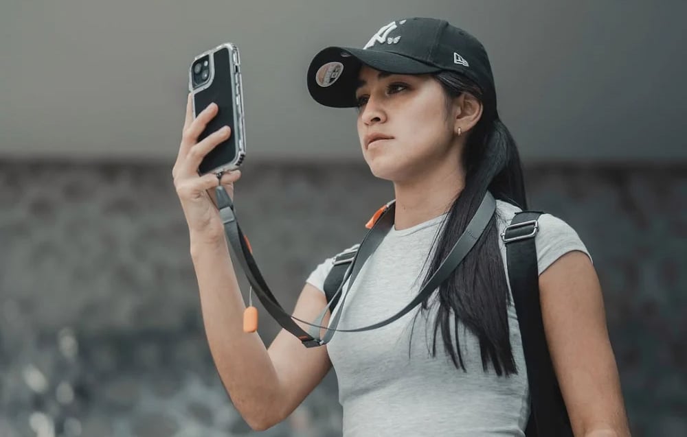 A woman wearing a baseball cap uses her phone, which is mounted to a gray neck strap
