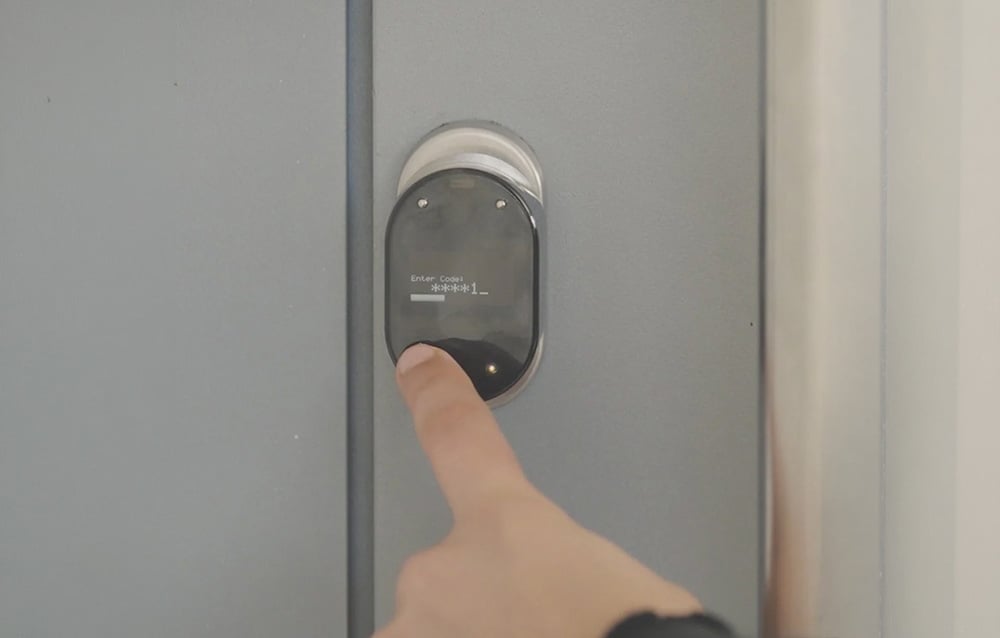 A finger presses a small, oval device with a display showing the code being entered on a gray door