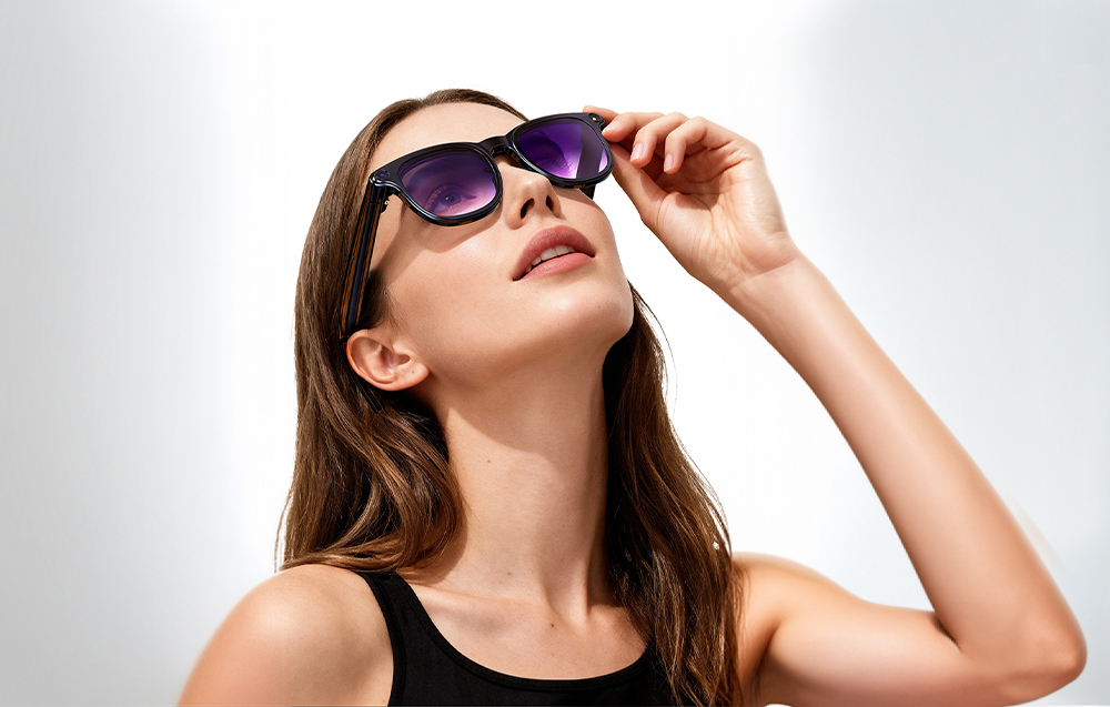 A woman in a black top adjusts her smart glasses with purple lenses while looking up