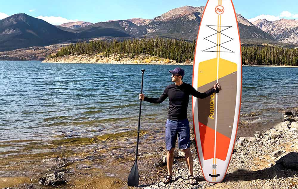 Man in a baseball cap standing on the shore of a lake holding a paddle and a surfboard, mountains in the background