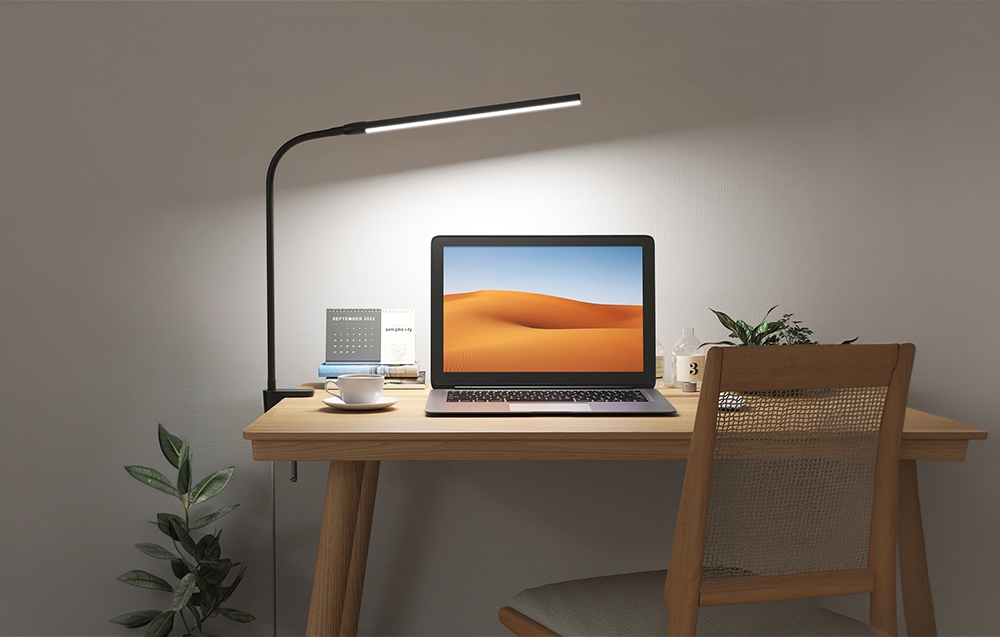 An illuminated workspace with a laptop, books, and a plant. A clamp lamp is attached to the side of a wooden desk.