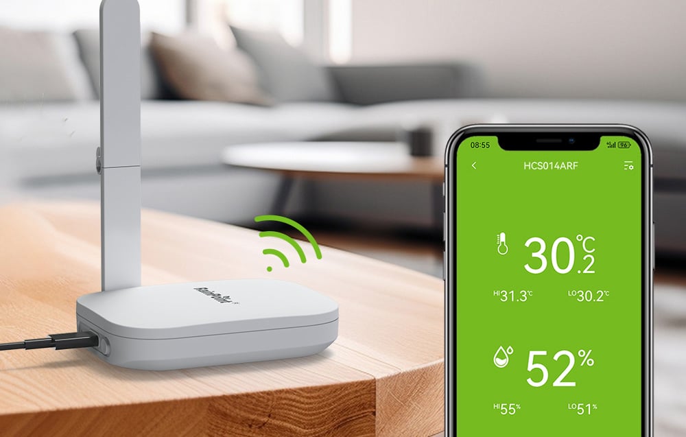 A white base station with an antenna sits on a wooden table next to a smartphone displaying a temperature of 30.2°C and humidity of 52%