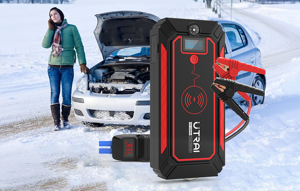 A car jump starter with jumper cables, with a woman standing next to a broken-down car in winter in the background.