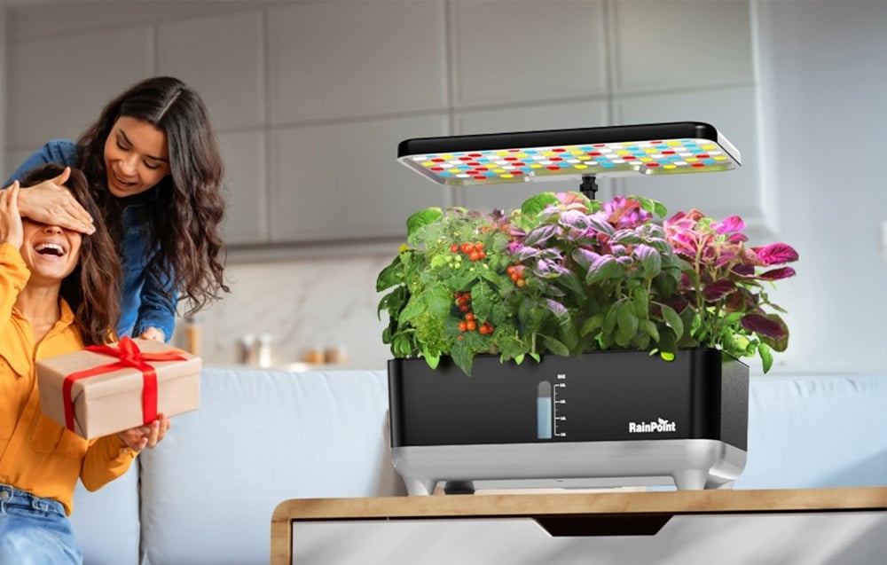 On the table next to two women and a gift stands a hydroponic system with a colorful LED panel turned on