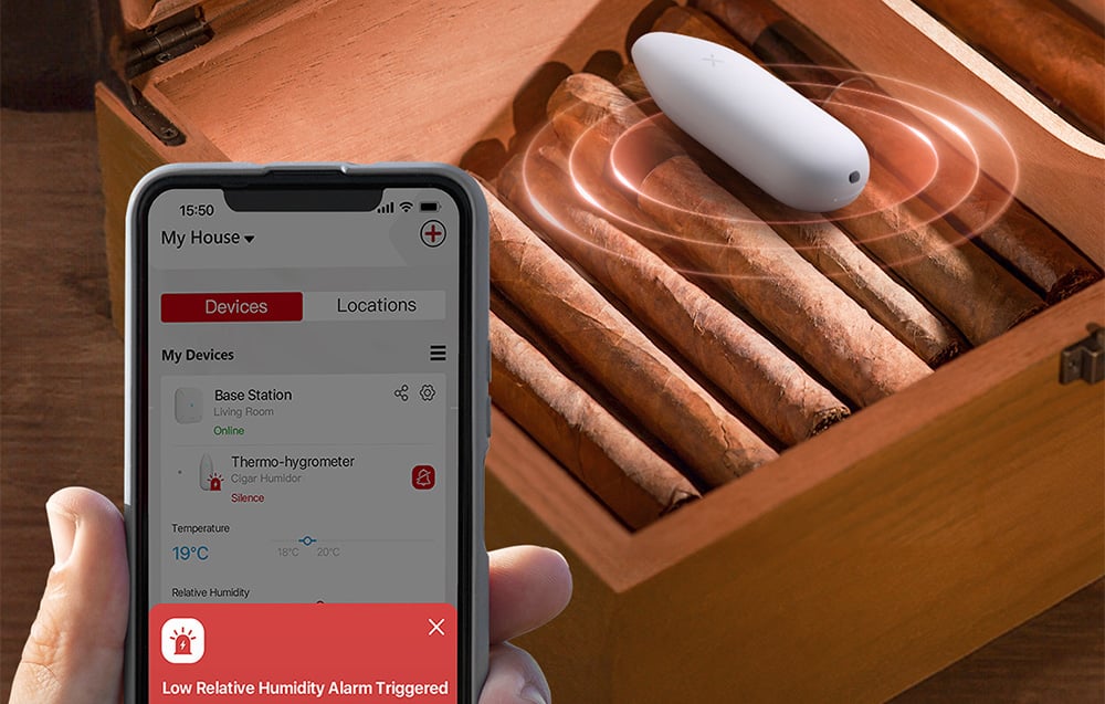 Telefon z aplikacją pokazującą alarm niskiej wilgotności przy drewnianej szkatułce z cygarami i białym czujnikiem