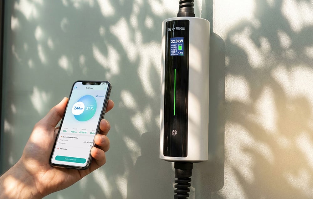 A smartphone held in the hand with the app open, an EV charger on the wall next to it