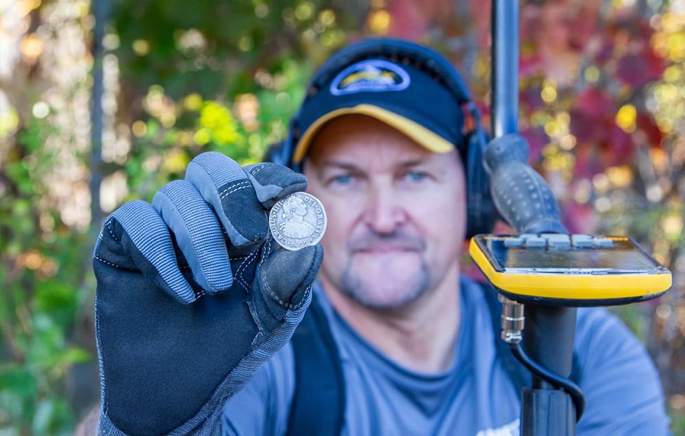 A man with a gloved hand holds a found coin, with a metal detector visible next to it.