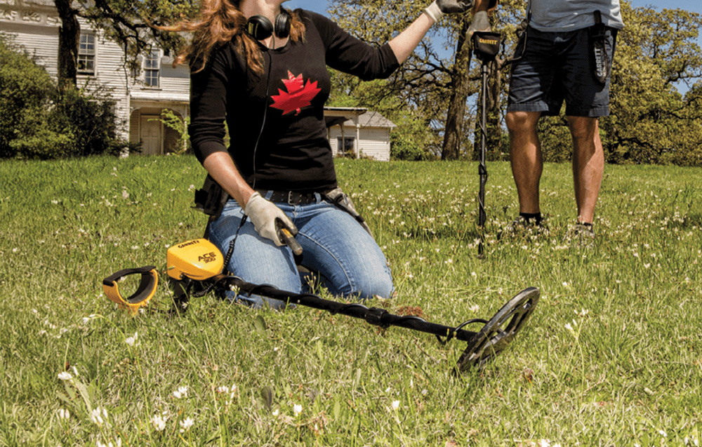 A woman kneels on the grass with a metal detector, a man stands next to her.