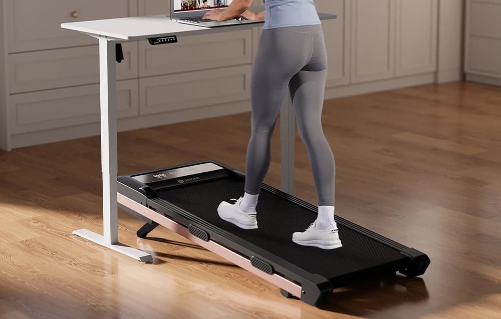 A person in gray leggings works at a white desk while standing on a black and pink treadmill
