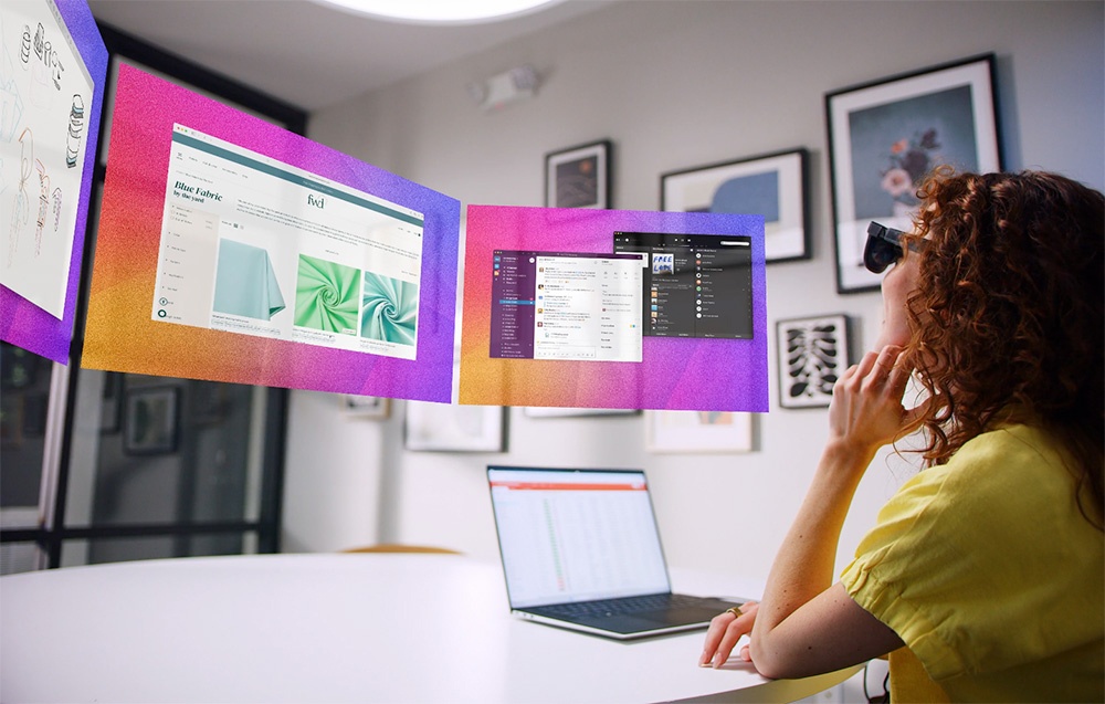 A woman in XR glasses works at a desk with a laptop, looking at virtual screens in the air