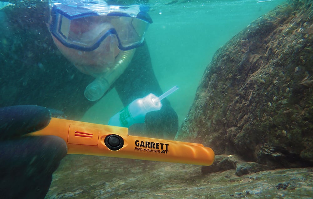 An underwater diver holds the metal detector against the rocks.