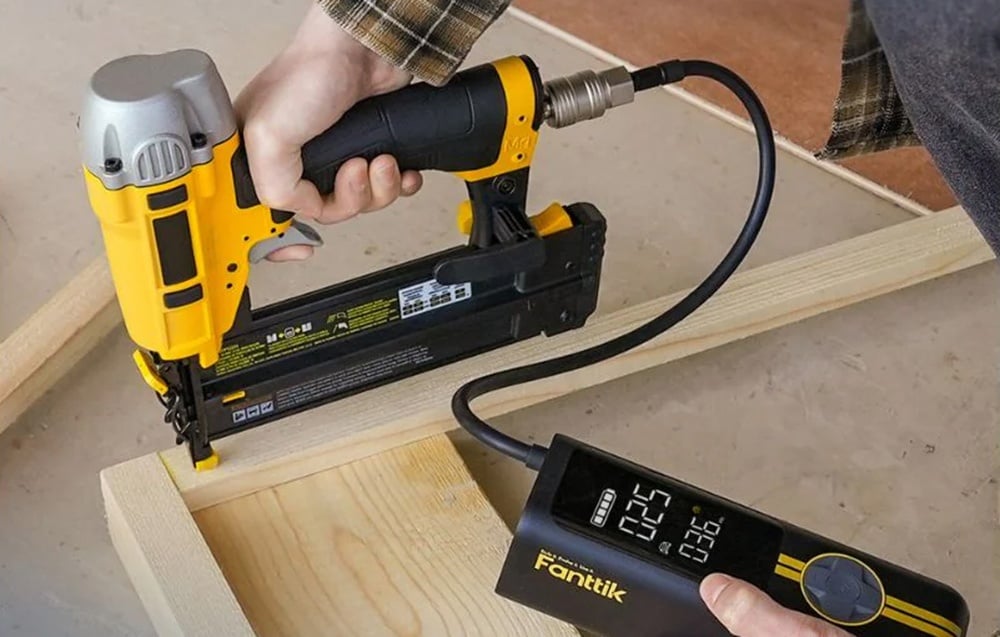A person uses a yellow and black pneumatic nailer connected to a portable compressor with a digital display, attaching wooden parts.