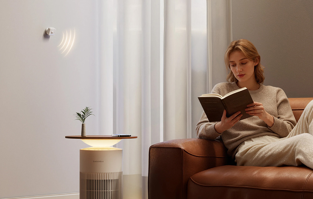 A presence sensor on the wall directs a signal toward a woman reading a book on the couch.