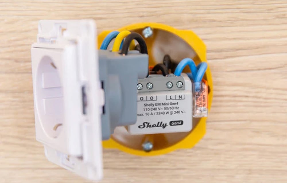 A white energy meter mounted inside a yellow electrical box in the wall, connected to an outlet with cables.