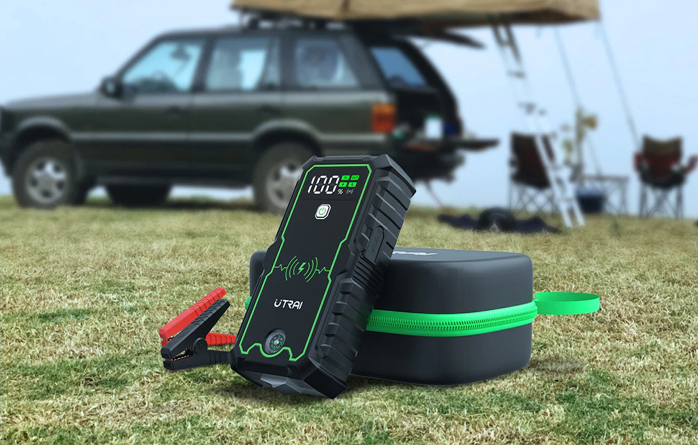 Portable jump starter with clamps and a carrying case, set against the backdrop of a campsite with a car.