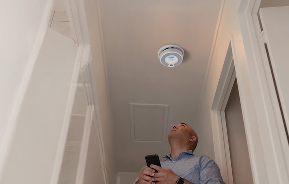 A man standing in the hallway under the ceiling looks at the installed smart smoke and carbon monoxide sensor