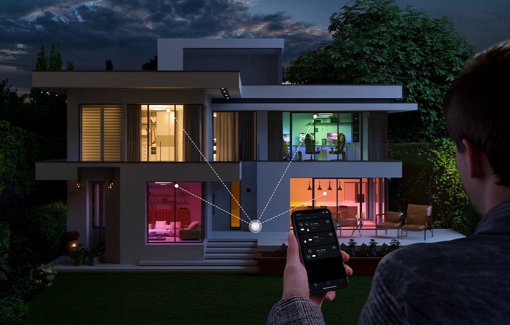 A two-story house at night with visible room lighting, controlled via a smartphone app.