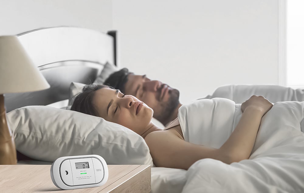 Para śpiąca w łóżku z białym detektorem CO na drewnianej szafce nocnej pokazującym 0 PPM