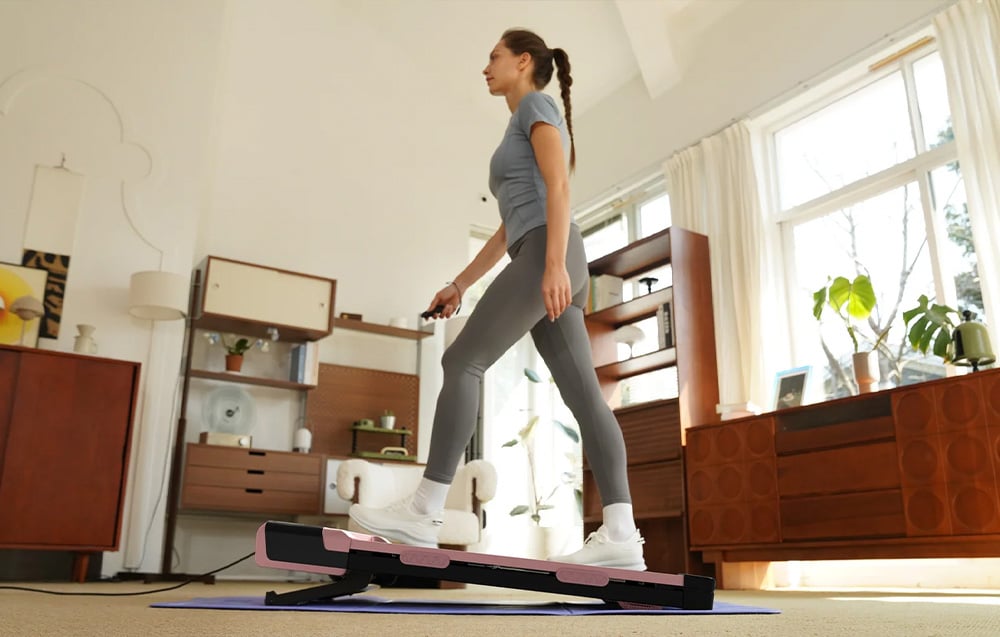 A woman walks on a compact pink and black treadmill in a bright living room with vintage furniture