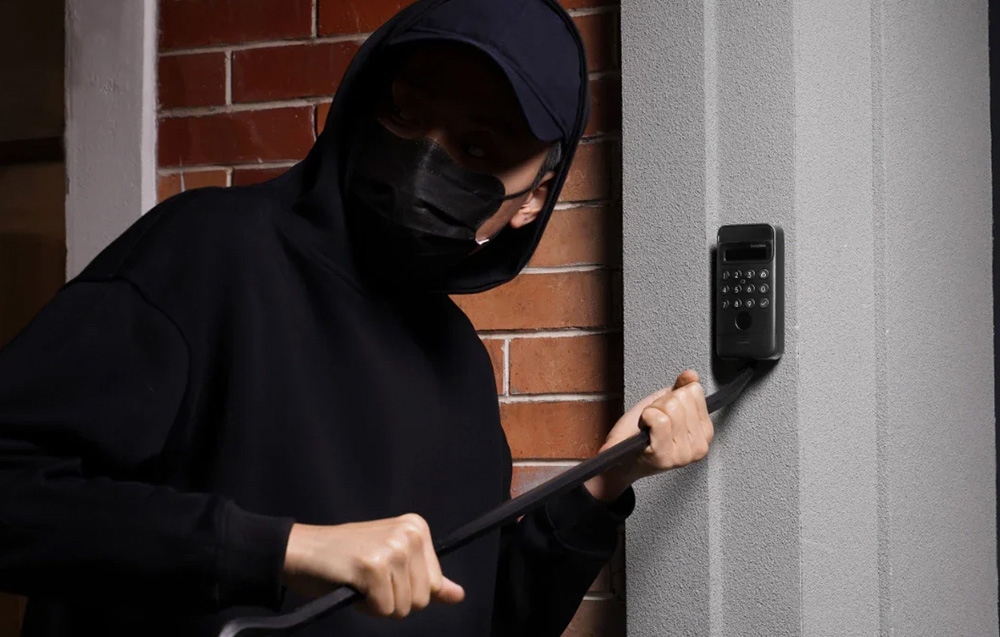 A burglar in a black sweatshirt with a chimney and glasses tries toлом the lock with a crowbar against a brick wall