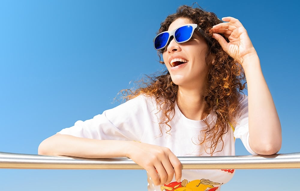 A woman wearing glasses with blue lenses leans against a metal railing against the backdrop of a clear, blue sky