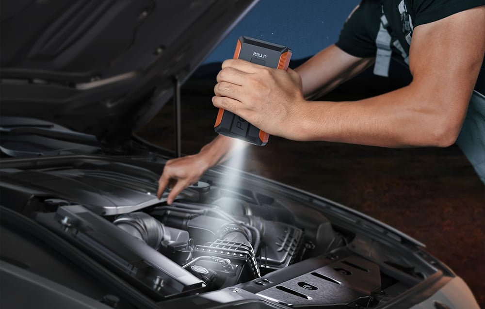 A man illuminates the interior of the open hood with a flashlight built into the jump starter.