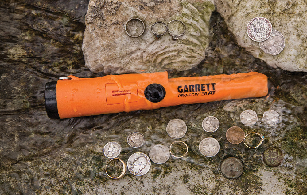 Handheld metal detector partially submerged in water with coins and rings.