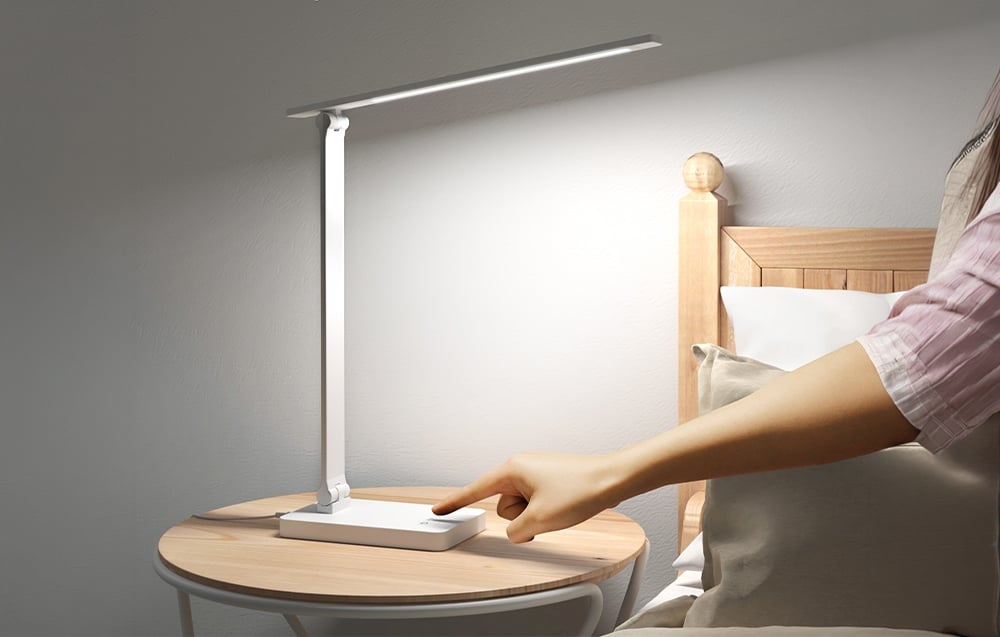 A hand touches the touch panel of a white table lamp standing on a round nightstand right next to a bed with pillows.