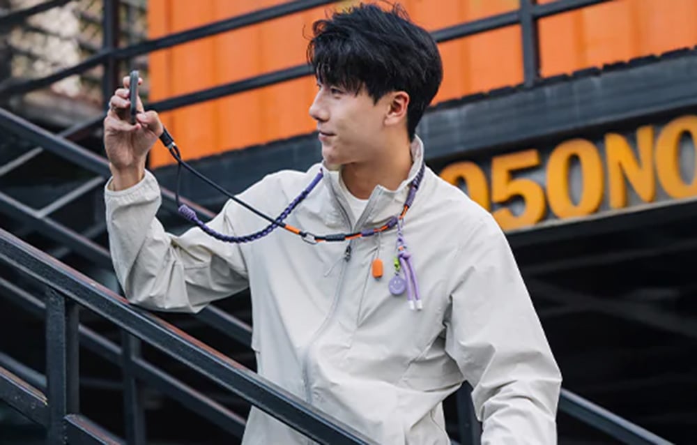 A man in a white jacket leans against the stairs and takes a photo with his smartphone, which is mounted to the PGYTECH strap