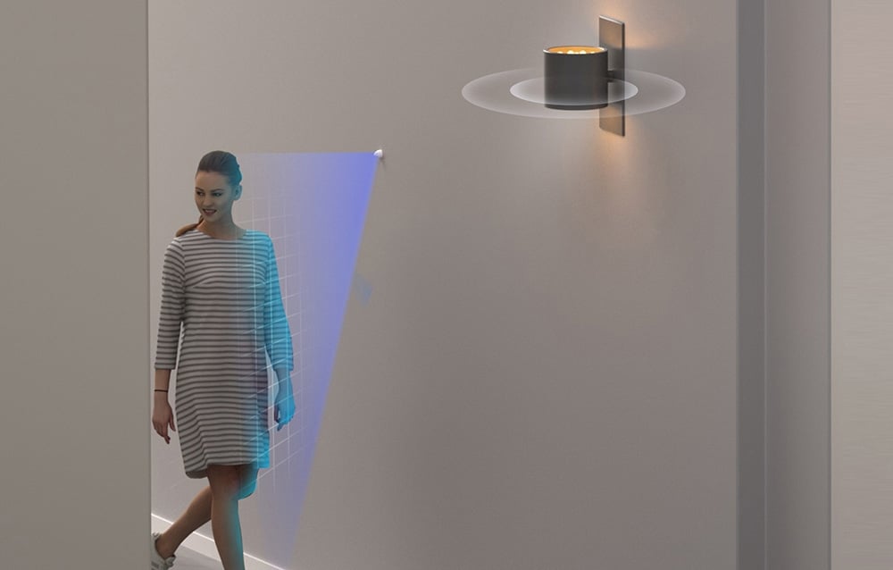 A woman walks past a motion sensor; a lamp is lit on the wall