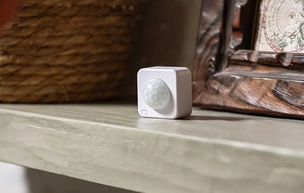 A small, white, square device with a round, convex lens featuring a grid pattern sits on a light-colored shelf