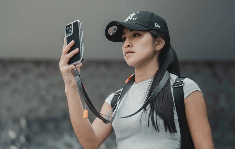 A woman wearing a baseball cap using her phone, which is attached to a neck strap