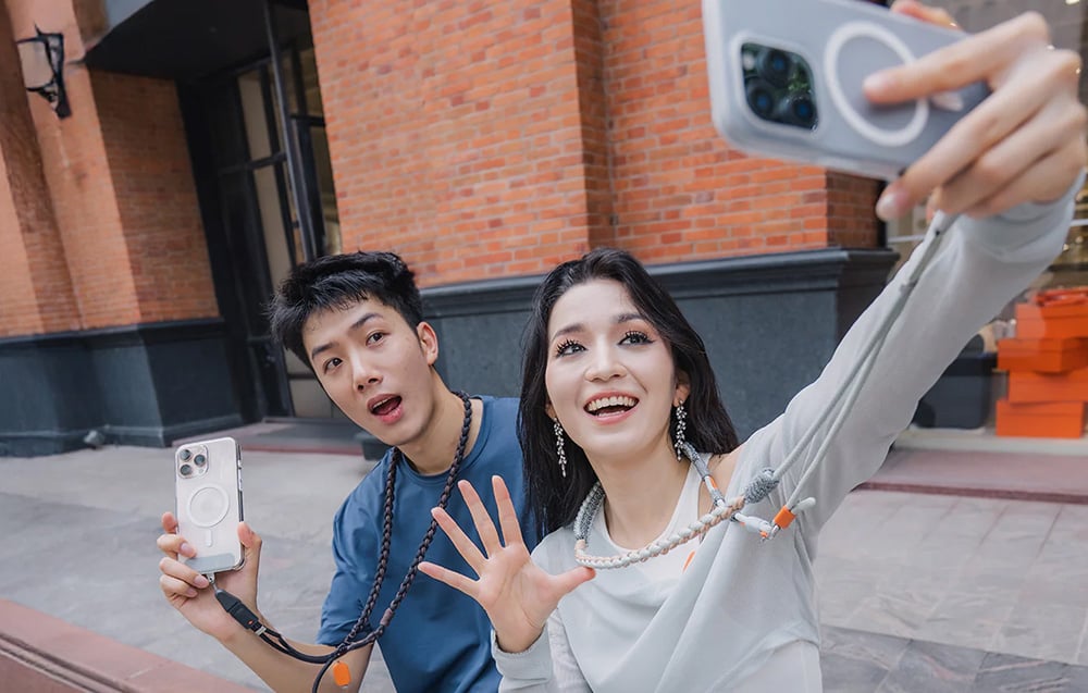 A woman and a man take a selfie against the backdrop of a brick building, both wearing a PGYTECH neck strap