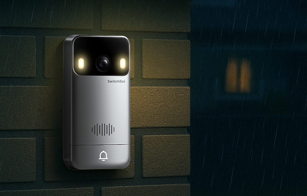 A silver doorbell with two glowing lights mounted on the building’s wall during a rainstorm.