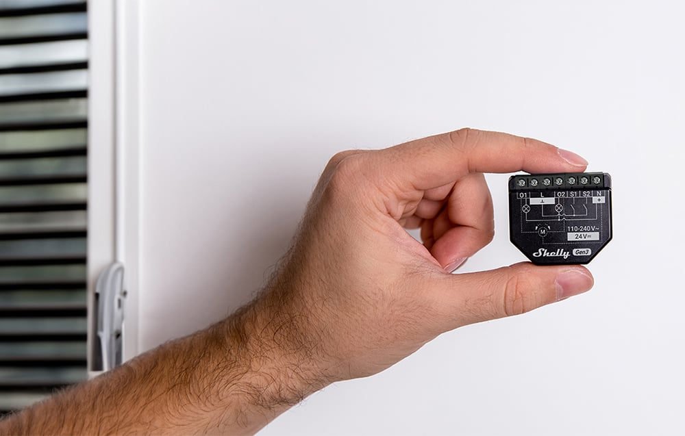 A hand holding a black Shelly 2PM Gen3 module against a white wall with a visible blind on the left side