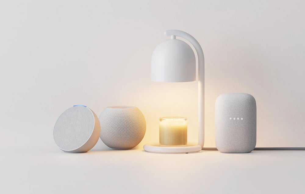A white candle warmer illuminating a glass scented candle, positioned between three smart home speakers: Amazon Echo Dot, Apple HomePod mini, and Google Nest Au