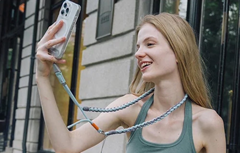 A blonde-haired woman takes a selfie with her smartphone connected to a PGYTECH strap