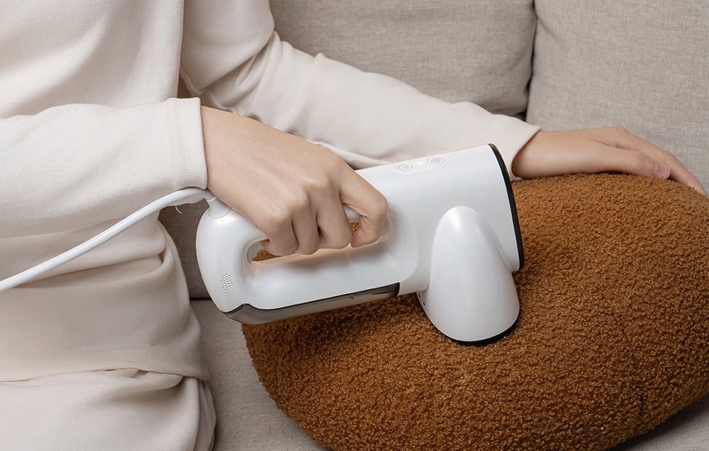 A person in a beige outfit sits on a couch and uses a white steamer to clean a brown cushion